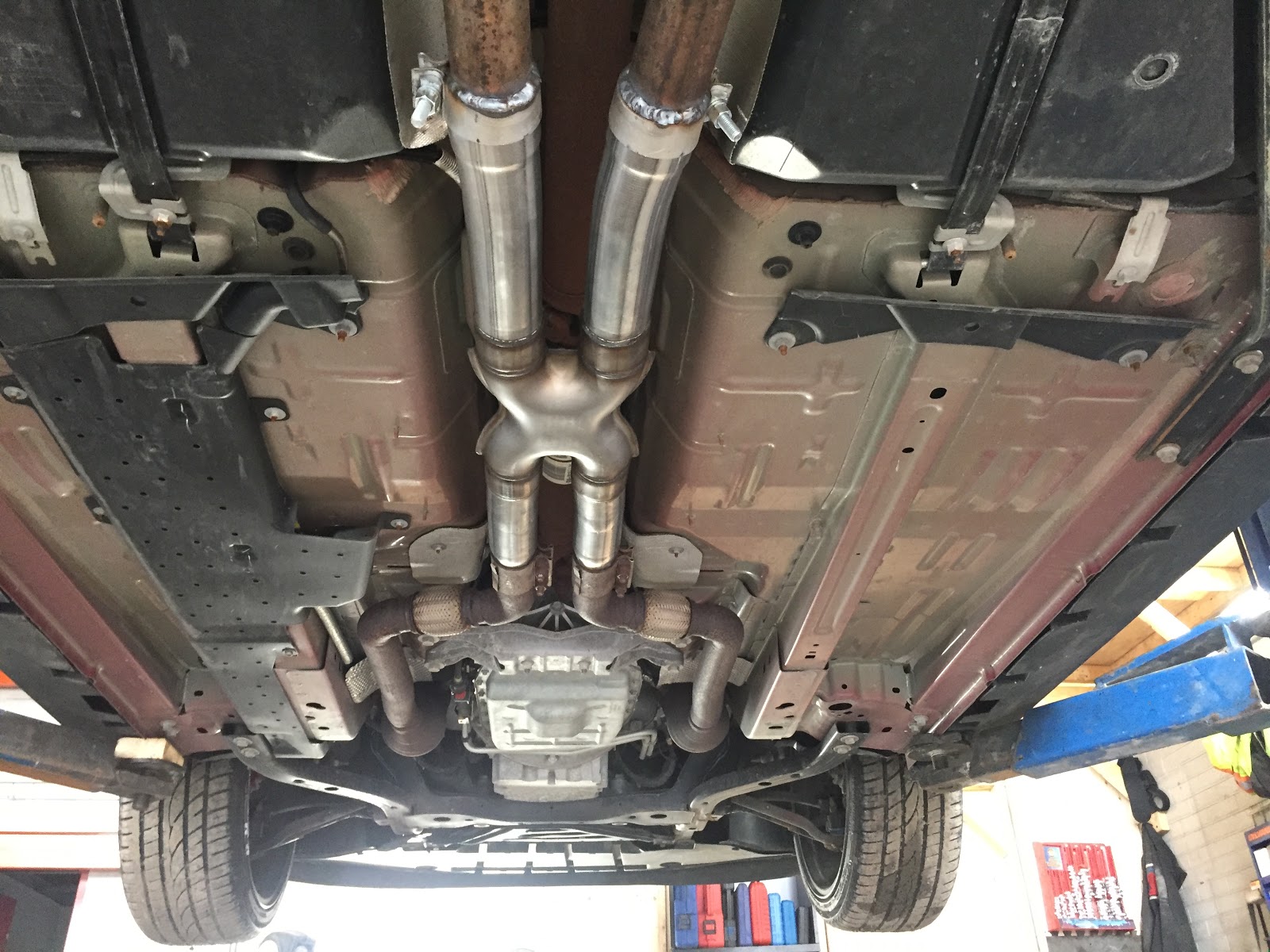 Underside of a car on the lift showing the X-pipe junction, headers, and exhaust routing in detail
